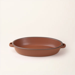 A large oval, matte cool terracotta ceramic baking dish with two side handles, on a clean, solid off-white background.