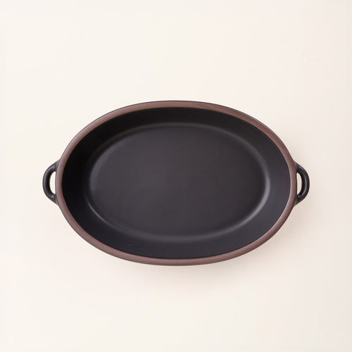 A large, oval, matte, graphite black ceramic baking dish with two side handles, on a clean, solid off-white background.