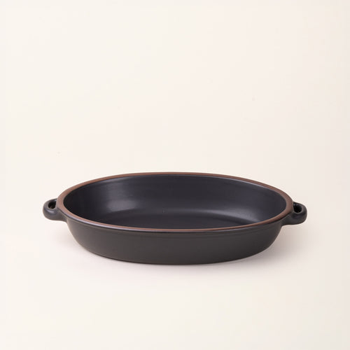 A large, oval, matte, graphite black ceramic baking dish with two side handles, on a clean, solid off-white background.