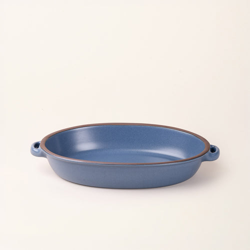 A large, oval, matte, grounded blue ceramic baking dish with two side handles, on a solid off-white background.