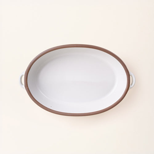 A large, oval, matte, cool white ceramic baking dish with two side handles, on a solid off-white background, photographed from overhead. 
