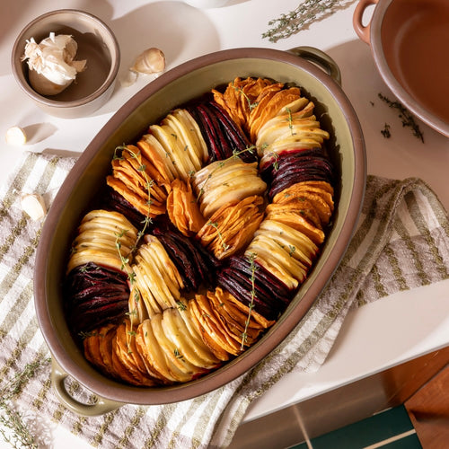 An overhead view of a large oval, olive green, ceramic baking dish with handles, filled with a root vegetable tian, slices stacked vertically inside the dish. The dish is surrounded by ceramic ramekins and sitting on a striped dish towel.