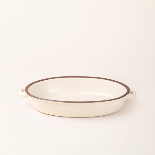 A large, oval, matte, warm off-white ceramic baking dish with two side handles, on a solid off-white background, photographed from overhead. 