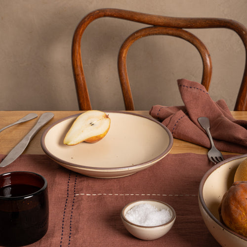 A medium ceramic dinner plate with a curved edge in a cream color, styled on a wooden table with a pear on top, alongside a brown linen napkin, cutlery, a small bowl of salt, and other tableware.