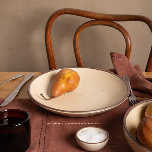 A large ceramic plate with a curved bowl edge in a matte cream color  on a table with brown linens, steel flatware and a small dark cup. A wooden chair back in the background. 