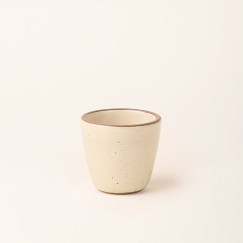 A small ceramic cup in a matte cream color featuring iron speckles, an unglazed rim and bottom base.