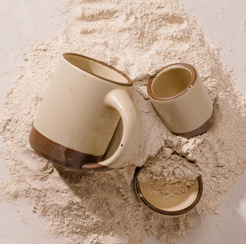 One cream colored ceramic mug, a small handle-less cup, and a small bowl arranged on a bed of light beige flour against a neutral background.