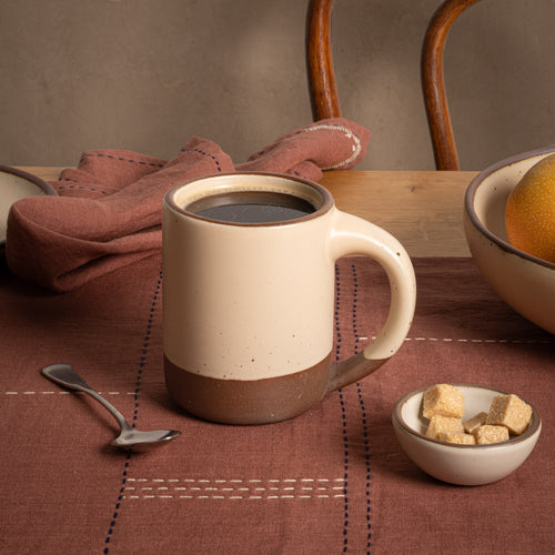 A medium sized ceramic mug with handle in a matte cream color featuring iron speckles, an unglazed rim and bottom base, filled with coffee on a table with brown linens and a small bowl of sugar cubes. 