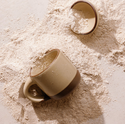 A small ceramic mug with handle in a matte cream color featuring iron speckles, an unglazed rim and bottom base placed on a surface surrounded by spilled flour and another small ceramic bowl in the same color.