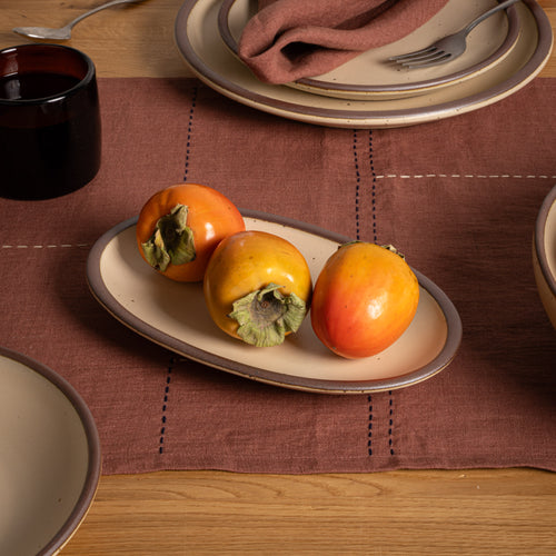 A small oval cream colored ceramic serving platter with an unglazed rim, styled on a brown linen placemat with three persimmon fruits, set on a wooden dining table alongside other plates and a dark glass cup.