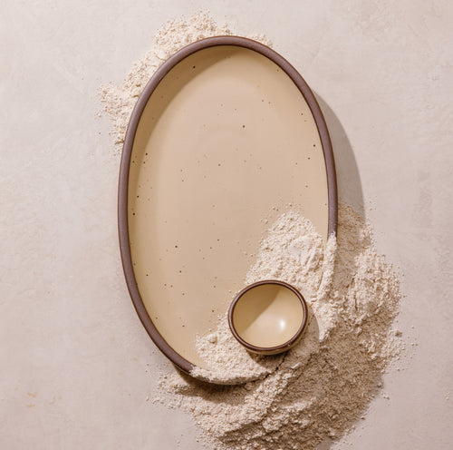 An overhead view of a large Oval shaped platter and tiny ceramic bowl in a matte cream color featuring iron speckles and unglazed rims, surrounded by scattered flour.
