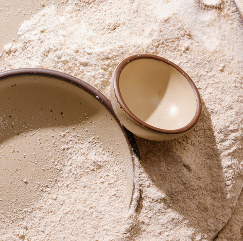 One matte cream colored, tiny ceramic bowl and one small plate filled with flour and arranged on a bed of loose flour.