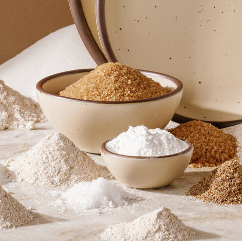 A styled product shot of various food powders, including white flour, white sugar, brown sugar, and baking powder, arranged in and around speckled cream colored ceramic bowls.