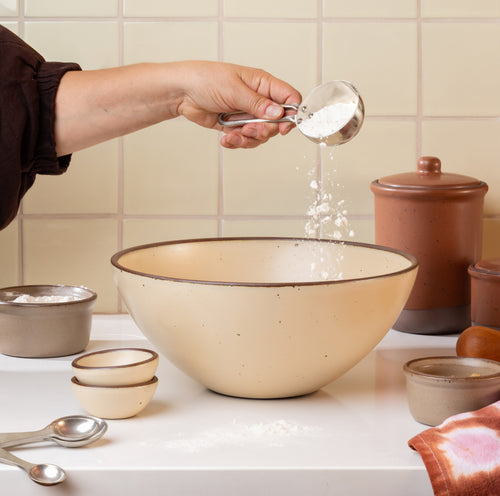 A person's hand pouring white flour from a metal measuring cup into a large cream colored ceramic mixing bowl with a dark rim, set on a white countertop with other ceramic bowls and baking items in a tiled kitchen setting.
