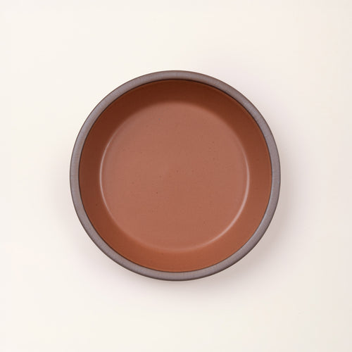 An overhead view of a shallow round ceramic pie plate in a terracotta color featuring an unglazed rim, against a cream background.