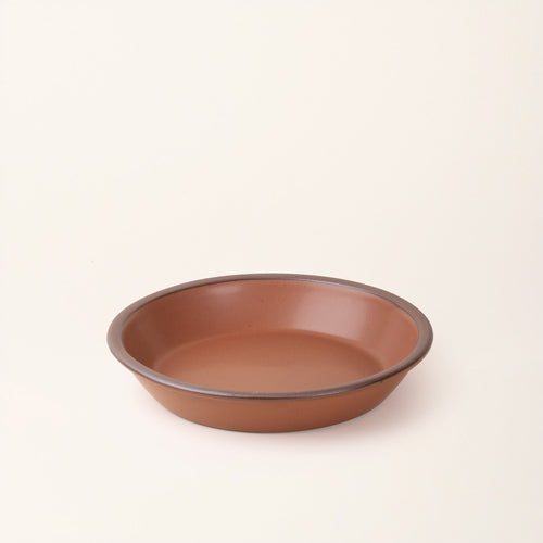 A shallow round ceramic pie plate in a matte terracotta color featuring an unglazed rim, photographed against a cream background.