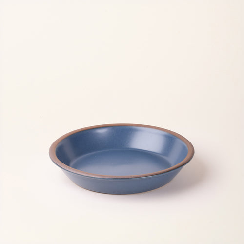 A shallow round ceramic pie plate in a mattegrounded blue color featuring an unglazed rim, photographed against a cream background.