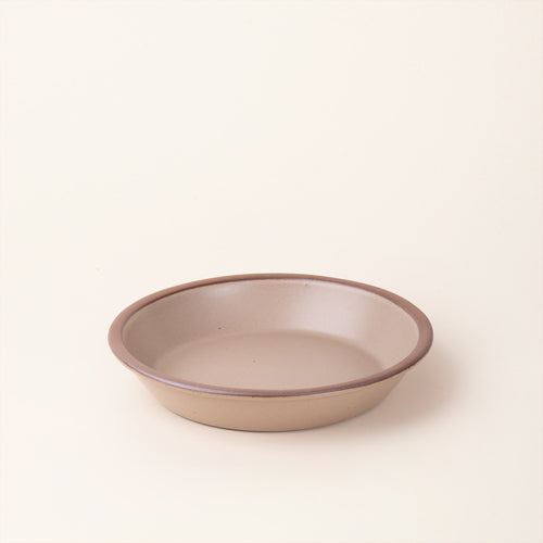 A shallow round ceramic pie plate in a matte pale warm brown color featuring an unglazed rim, photographed against a cream background.
