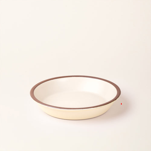A shallow round ceramic pie plate in a matte warm off-white color featuring an unglazed rim, photographed against a cream background.
