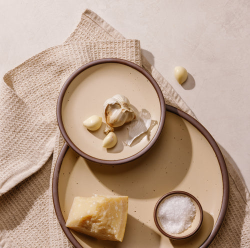 A styled flat-lay of matte cream colored ceramic serving plates on a textured beige cloth. A small plate holds a garlic bulb and cloves, while a medium below holds a wedge of hard cheese and a small bowl of coarse salt. 