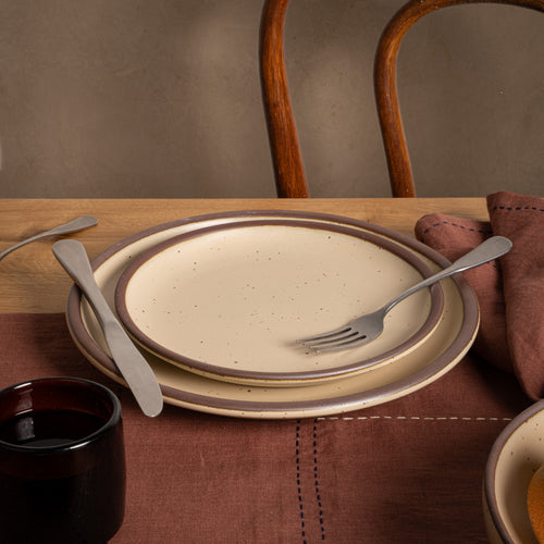 A medium sized ceramic plate in a matte cream color featuring iron speckles and an unglazed rim, stacked ontop of a dinner plate. Steel cutlery, a dark glass with liquid join the plates on a brown placemat. 
