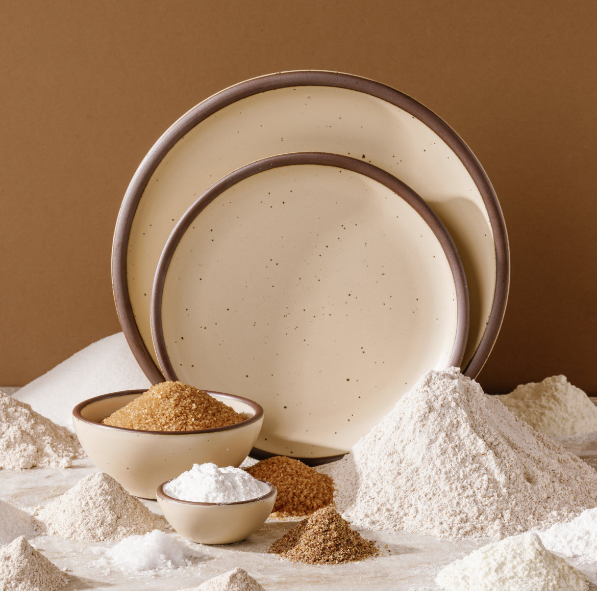 Two round ceramic plates  that are a matte cream color styled with bowls of flour and baking ingredients on a brown background.