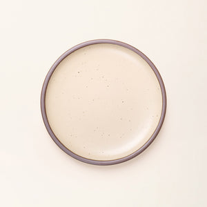 A dinner sized ceramic plate in a matte cream color featuring iron speckles and an unglazed rim.