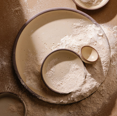 An overhead view of a large shallow ceramic bowl, a medium bowl, and a small cup in a matte cream color featuring iron speckles and unglazed rims, surrounded by scattered flour.