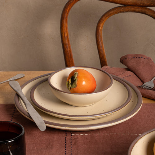A set of ceramic tableware including a small matte cream-colored bowl with a brown rim, a matching plate, and a larger plate, styled on a table with a brown placemat, silver cutlery, a folded brown napkin, and a persimmon in the center bowl.