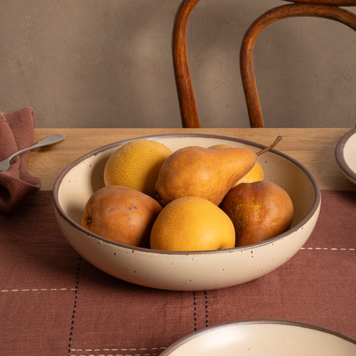 A dinner sized ceramaic bowl in a cream color filled with pears, set on a rustic wooden table with a brown placemat and chair in the background.