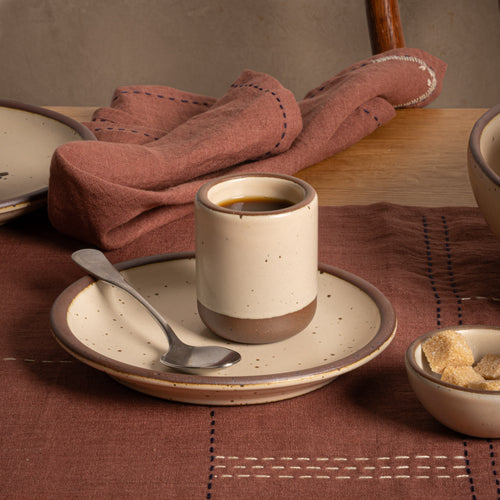 A tiny ceramic cup in a matte cream color with coffee inside, sitting on a small saucer with a spoon and raw sugar cubes nearby. 