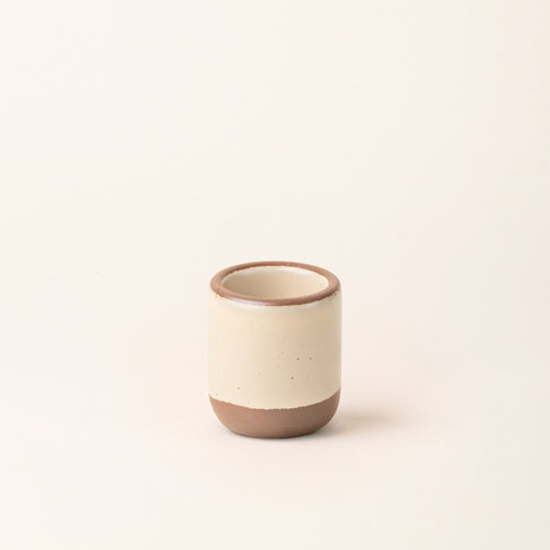 A tiny ceramic cup in a matte cream color featuring iron speckles, an unglazed rim and bottom base.