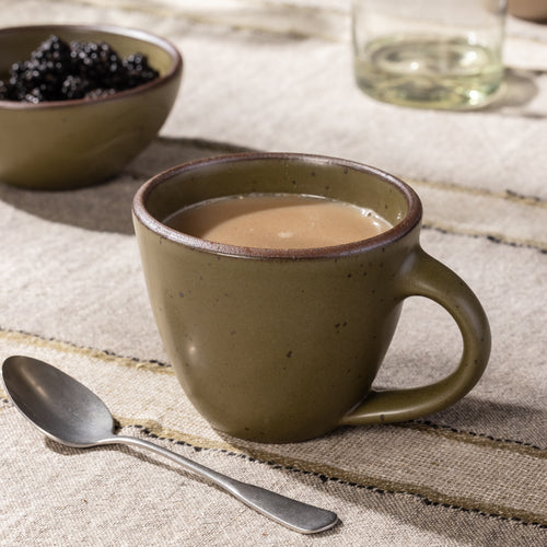 Olive-glazed stoneware mug with a rounded silhouette and speckled finish, styled with coffee on a rustic linen table.