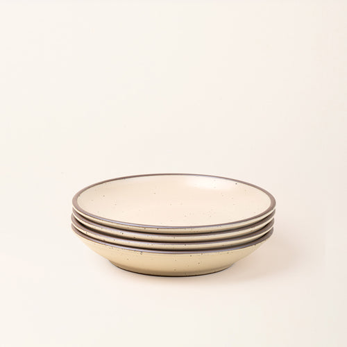 A stack of 4 large ceramic plates with a curved bowl edge in a matte cream color featuring iron speckles and unglazed rims.
