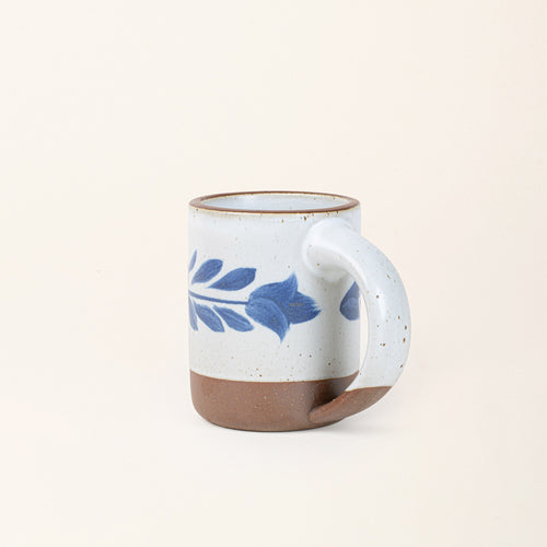 Side profile of a White ceramic mug with a hand-painted blue leaf motif and exposed clay base on a cream background.