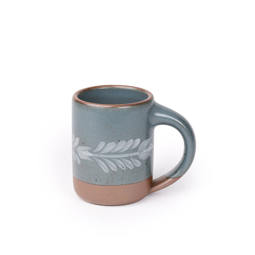 Blue-grey ceramic mug with a hand-painted white leaf motif and exposed clay base on a plain white background.