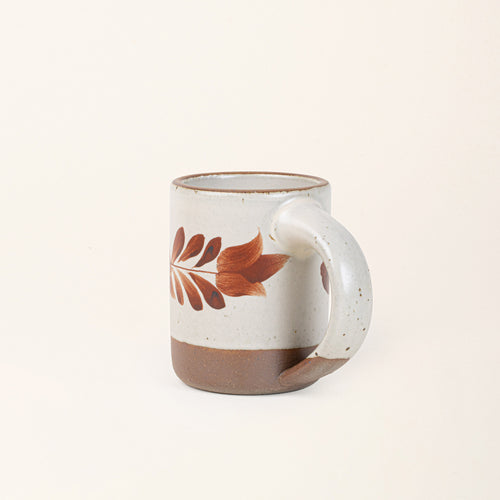 Side profile of a Warm off-white ceramic mug with a hand-painted terracotta leaf motif and exposed clay base on a cream background.
