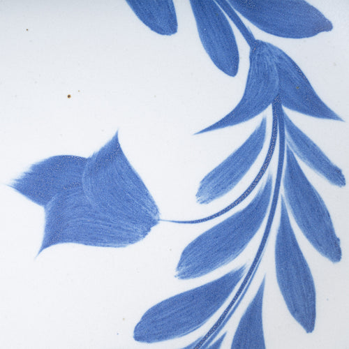 Closeup of an Oval ceramic platter with a hand-painted blue leaf and flower border on a white background with an exposed clay rim.
