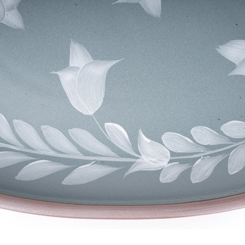 Closeup of an Oval ceramic platter with a hand-painted white leaf and flower border on a blue-grey background with an exposed clay rim.