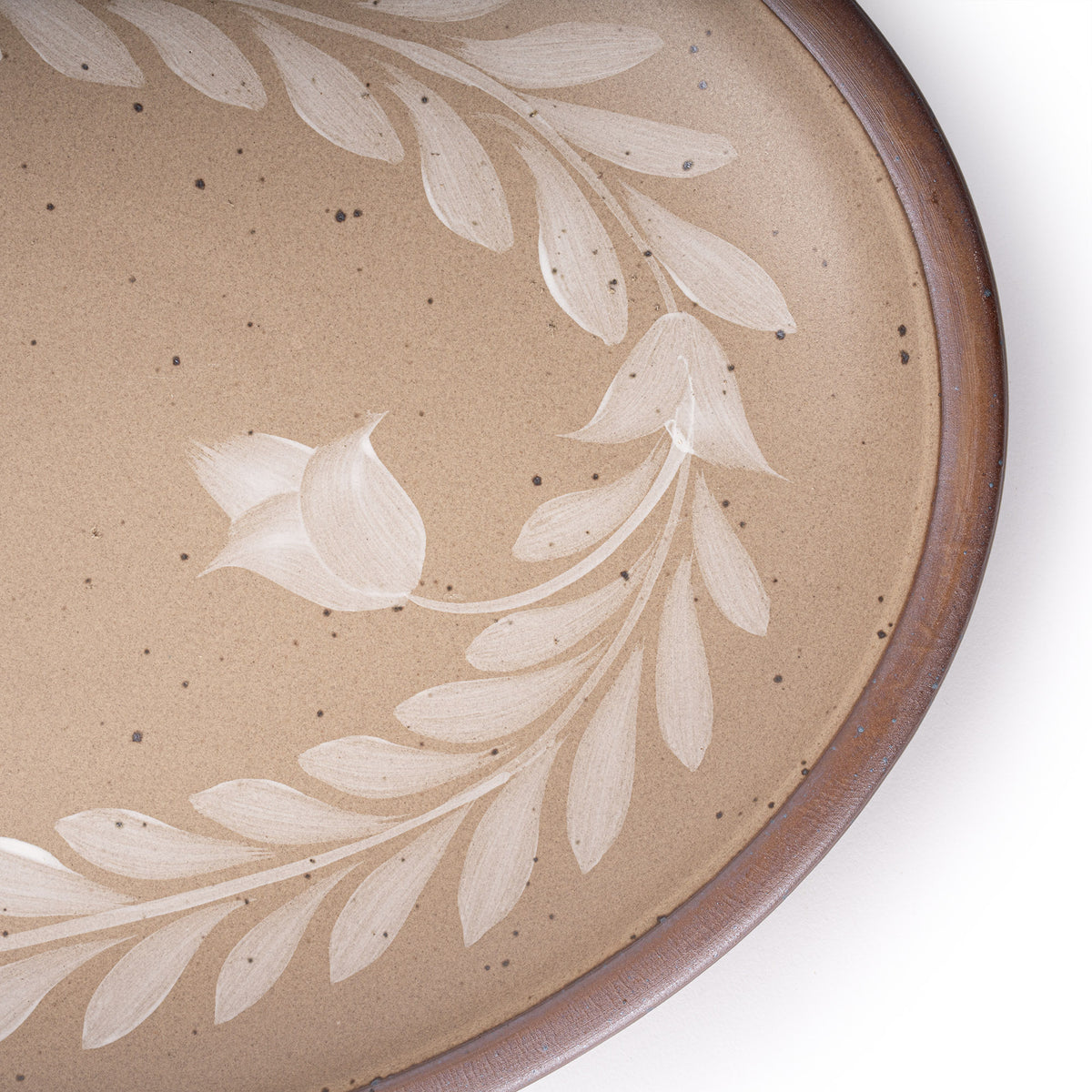 Close up of an Oval ceramic platter with a hand-painted white leaf and flower border on a warm pale brown background with an exposed clay rim.