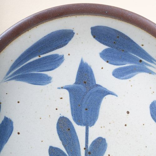 Closeup of a small round ceramic plate with a hand-painted blue flower and leaf design on a white background with an exposed clay rim.