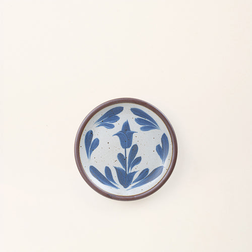 Small round ceramic plate with a hand-painted blue flower and leaf design on a white background with an exposed clay rim.