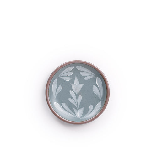 Small round ceramic plate with a hand-painted white flower and leaf design on a blue-grey background with an exposed clay rim.