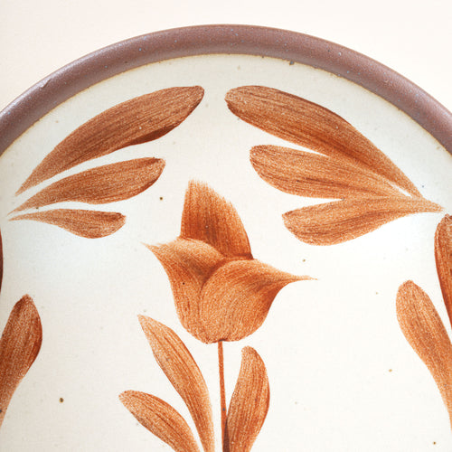 Closeup of a small round ceramic plate with a hand-painted terracotta flower and leaf design on a cream background with an exposed clay rim.