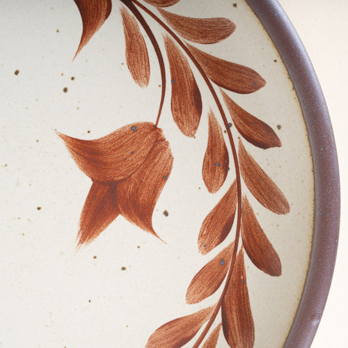 Closeup of a side round ceramic plate with a hand-painted terracotta leaf wreath and floral design on a cream background with an exposed clay rim.