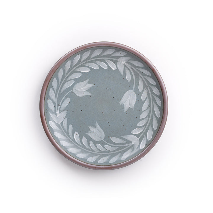 Dinner round ceramic plate with a hand-painted white leaf wreath and floral design on a blue-grey background with an exposed clay rim.
