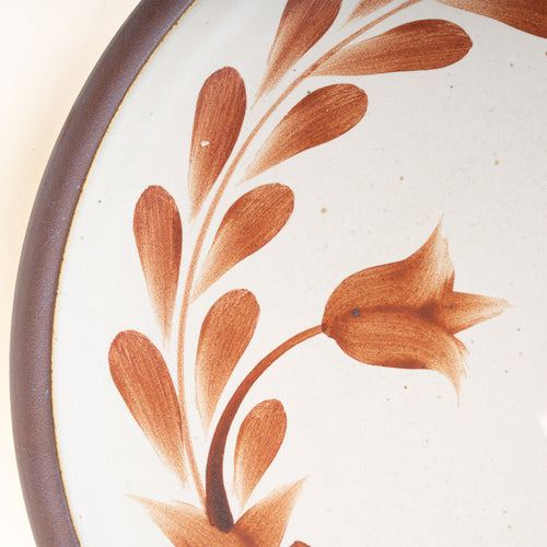 Closeup of a dinner round ceramic plate with a hand-painted cool terracotta leaf wreath and floral design on a cream background with an exposed clay rim.