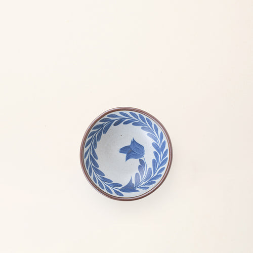 An overhead view of a Small ceramic shallow bowl with a hand-painted blue leaf border on a white interior and speckled white exterior with an exposed clay rim.