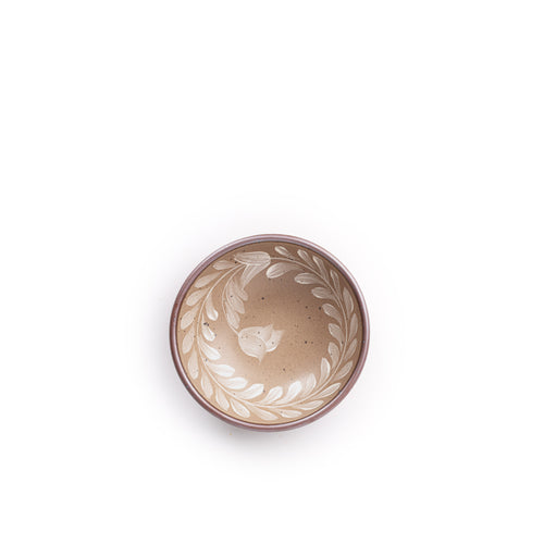 Overhead view of a small ceramic shallow bowl with a hand-painted white leaf border on a pale warm brown interior and exterior with an exposed clay rim.