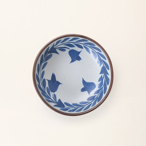 An overhead view of a medium ceramic shallow bowl with a hand-painted blue leaf border on a white interior and exterior with an exposed clay rim.